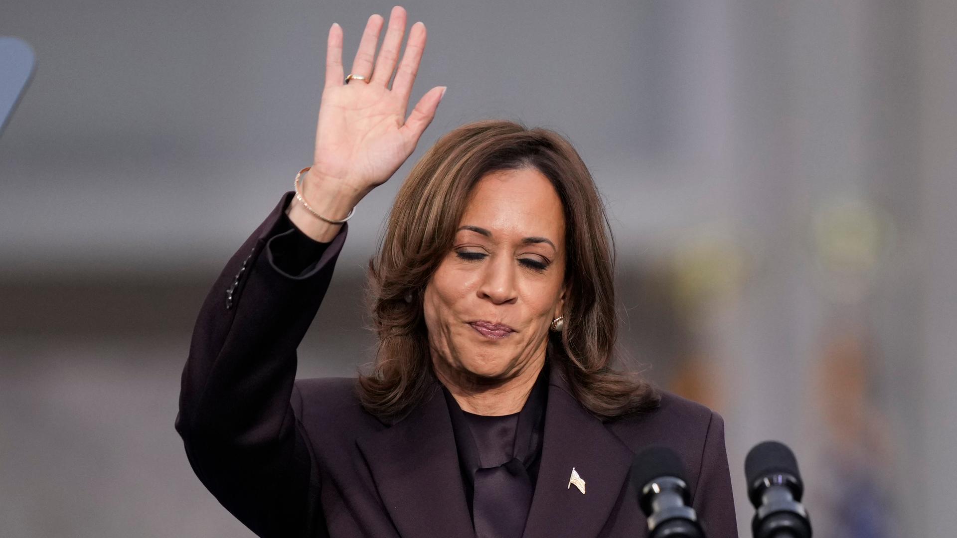 Kamala Harris steht hinter Mikrofonen und winkt. 