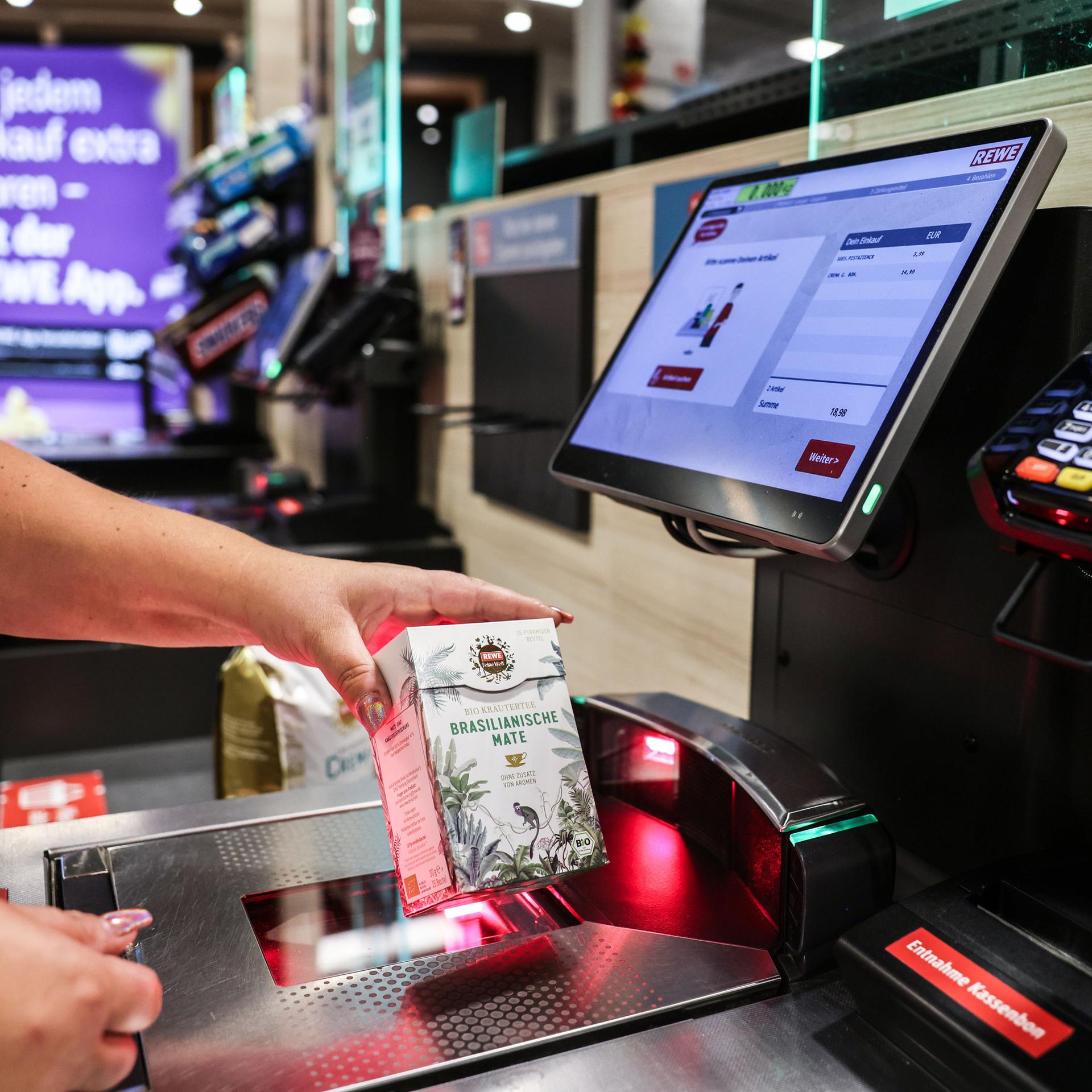 Eine Frau scannt in einer Rewe-Fliale ein Produkt an einer Selbstbedienungskasse. 
