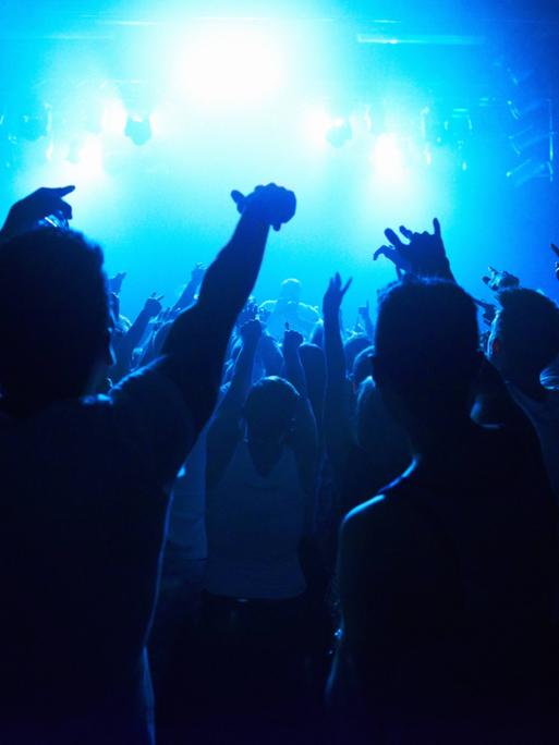 Menschen halten ihre Hände in einem Nachklub nach oben, um sie herum blaues Licht und Nebel