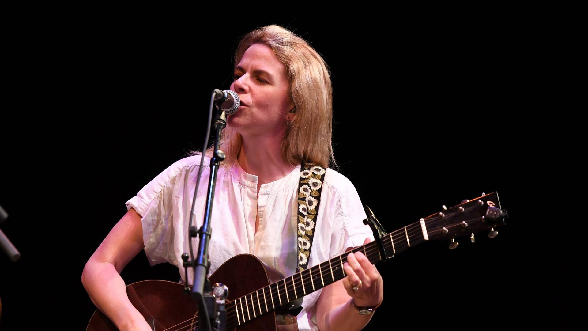Eine Sängerin mit schulterlangen, blonden Haaren steht unprätentiös auf einer Bühne und singt, während sie sich mit der Gitarre begleitet.