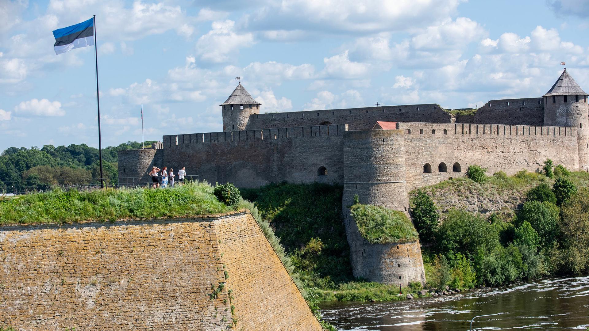 Die estnische Flagge weht über einer Gruppe von Personen, die über den Fluss Narva hinüber zur Festung Iwangorod auf der russischen Seite des Flusses schauen.