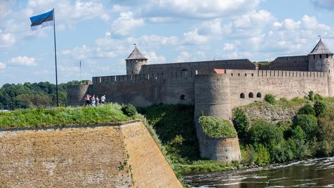 Die estnische Flagge weht über einer Gruppe von Personen, die über den Fluss Narva hinüber zur Festung Iwangorod auf der russischen Seite des Flusses schauen.
