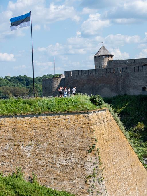 Die estnische Flagge weht über einer Gruppe von Personen, die über den Fluss Narva hinüber zur Festung Iwangorod auf der russischen Seite des Flusses schauen.