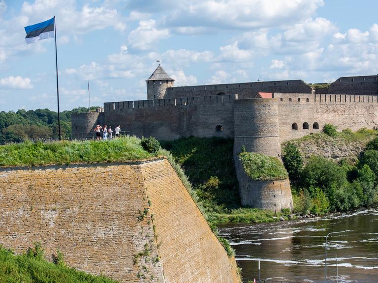 Die estnische Flagge weht über einer Gruppe von Personen, die über den Fluss Narva hinüber zur Festung Iwangorod auf der russischen Seite des Flusses schauen.