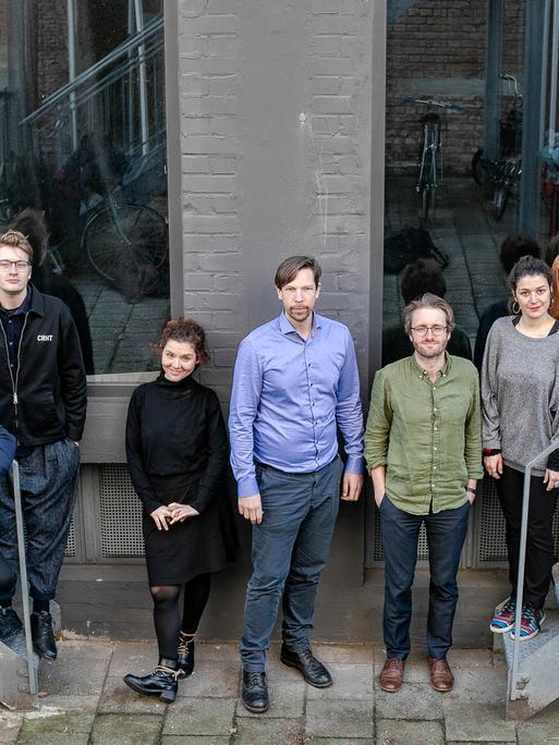 Eine Gruppe von acht Männern und zwei Frauen steht zwischen zwei Treppen vor einem Gebäude mit großen Fenstern in einer Reihe. 