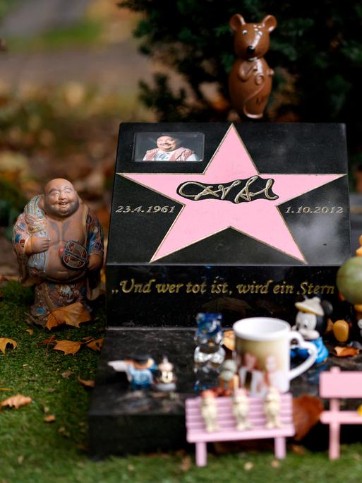 Das Grab von Entertainer Dirk Bach auf dem Melaten-Friedhof: Immer wieder bringen Fans kleine Figürchen vorbei oder haben auf einer rosa Bank am Grab eine 'Audienz beim Mausekönig'. Die Stadt Köln ließ alle Bänke entfernen.
