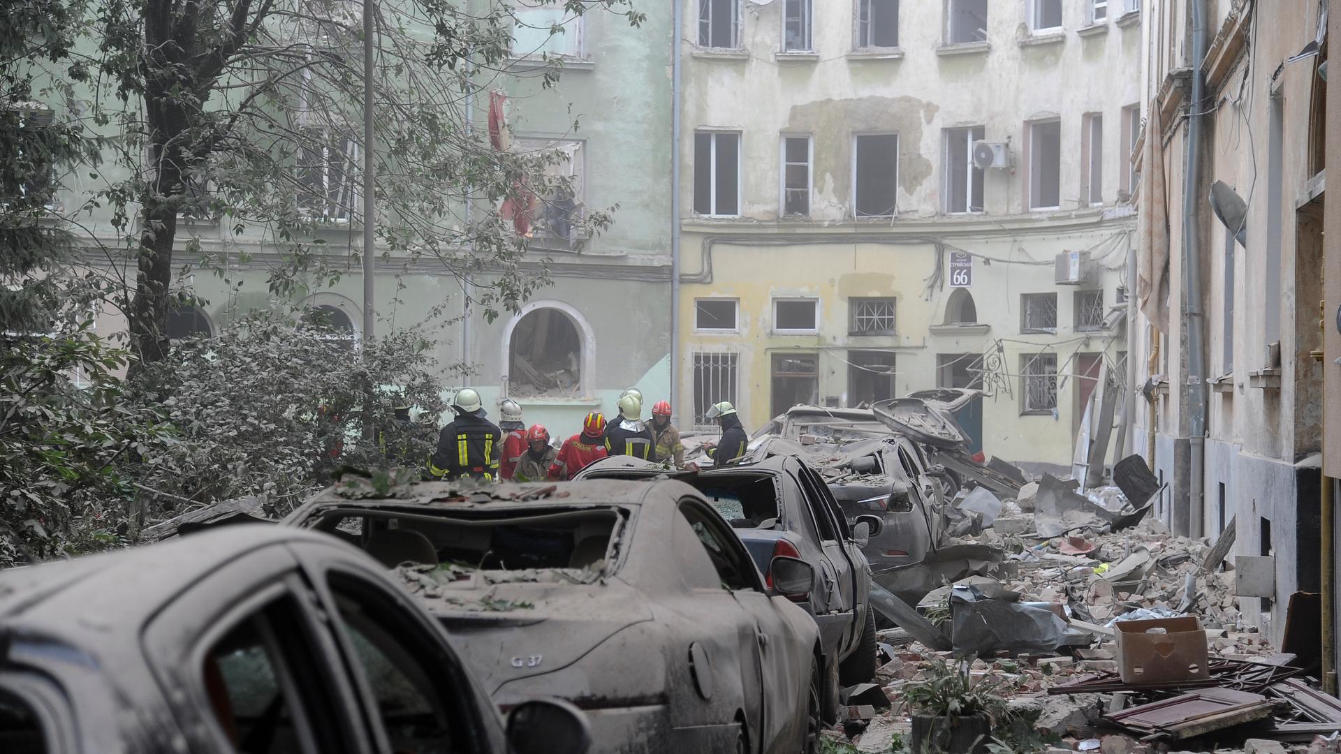 Rettungskräfte versammeln sich in Lwiw nach einem russischen Raketenangriff vor beschädigten Gebäuden. Im Vordergrund sind beschädigte Autos und Trümmer zu sehen.