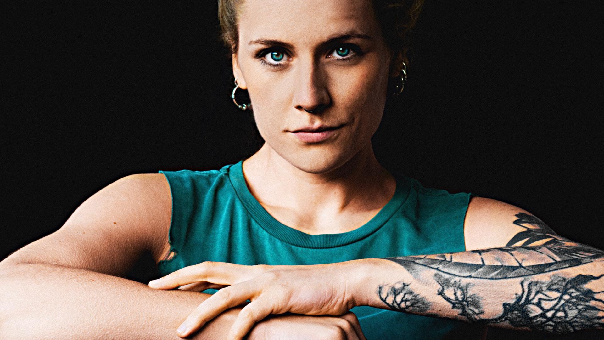 Laura Braun trägt ein schulterfreies blaues Top und verschränkt ihre Arme. Ihr rechter Arm ist tätowiert.