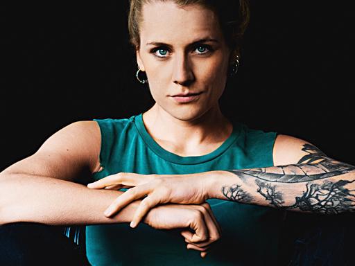 Laura Braun trägt ein schulterfreies blaues Top und verschränkt ihre Arme. Ihr rechter Arm ist tätowiert.