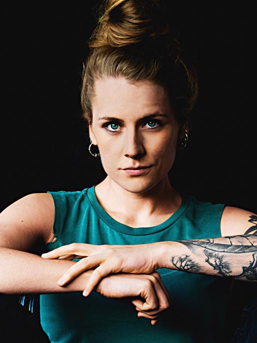 Laura Braun trägt ein schulterfreies blaues Top und verschränkt ihre Arme. Ihr rechter Arm ist tätowiert.