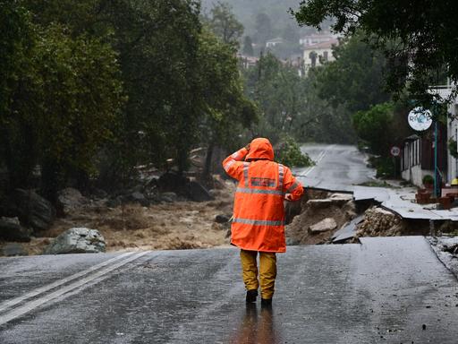 Eine Person in oranger Warnweste steht auf einer regennassen Straße, die durch Unterspülungen eingebrochen ist.