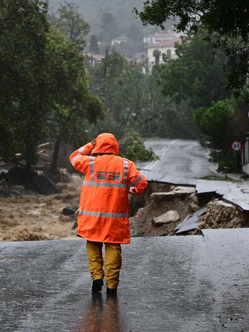 Eine Person in oranger Warnweste steht auf einer regennassen Straße, die durch Unterspülungen eingebrochen ist.