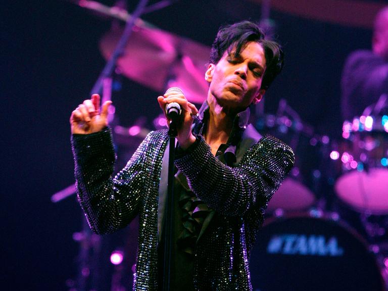 Der Sänger Prince mit geschlossen Augen und einem Mikrofon in der Hand beim Auftritt auf der Bühne.