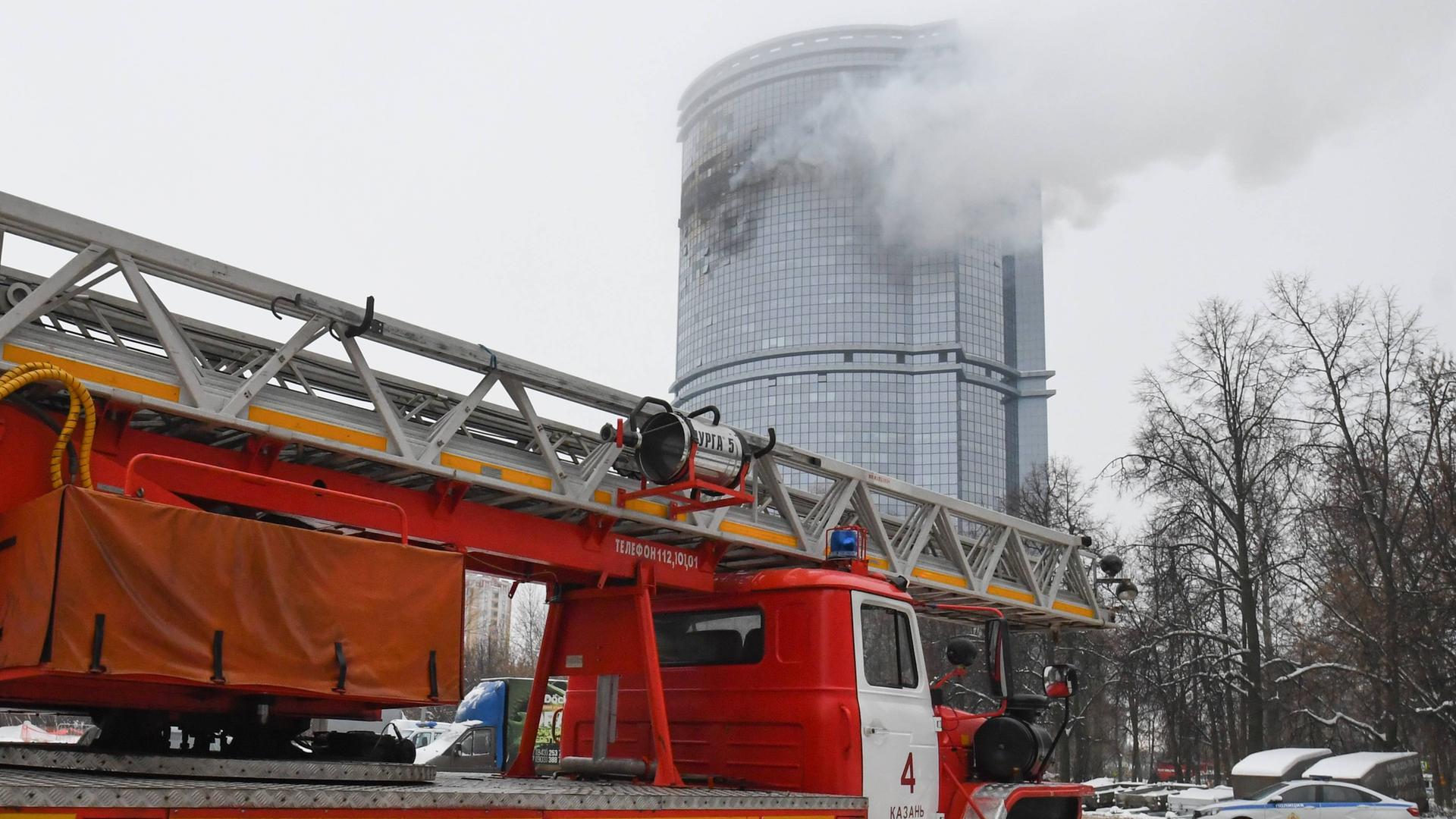 Zu sehen ist ein Feuerwehrauto, das vor einem Hochhaus steht. Aus höheren Stockwerken dringt durch ein Feuer verursachter Rauch nach außen. Zu sehen ist ein Feuerwehrauto, das vor einem Hochhaus steht. Aus höheren Stockwerken dringt durch ein Feuer verursachter Rauch nach außen.