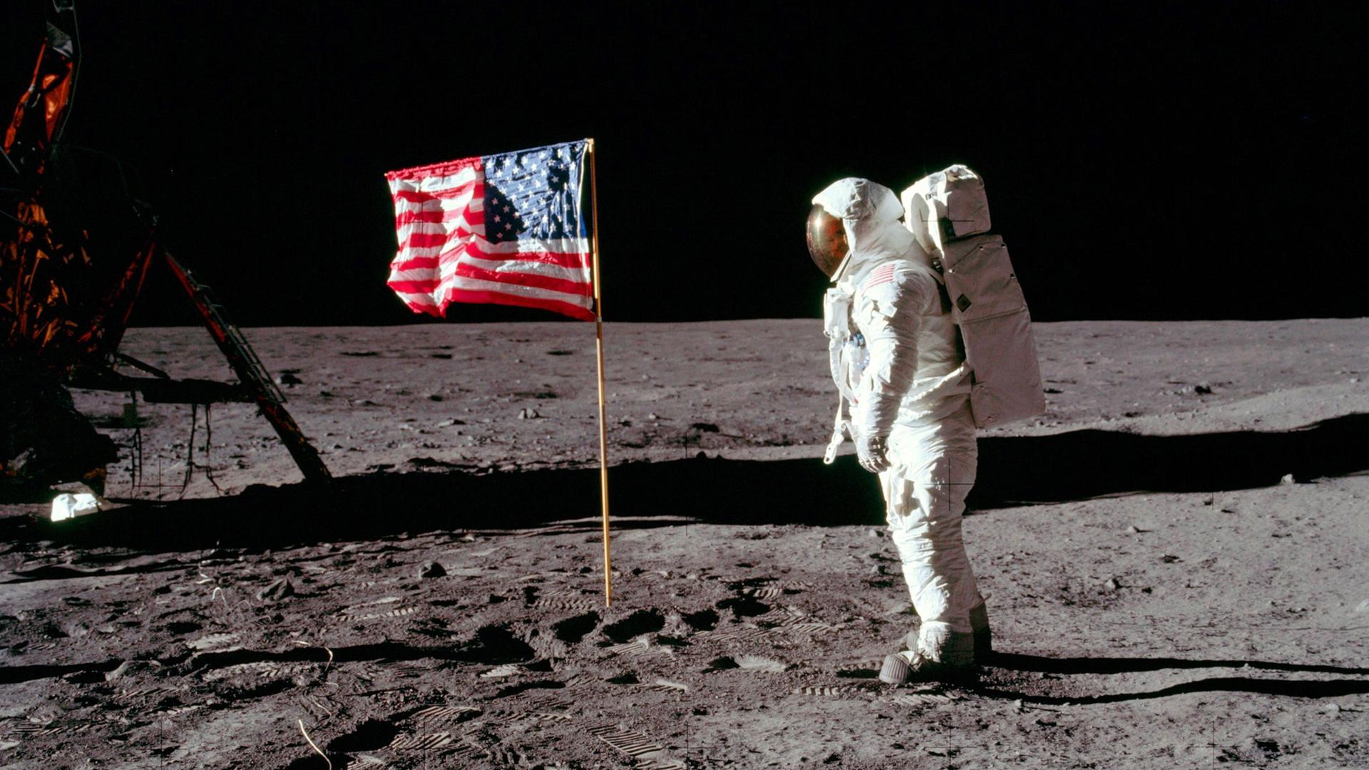 Historisches Foto, auf dem Neil Armstrong in seinem Raumanzug auf der Mondoberfläche steht und auf die US-amerikanische Flagge schaut.