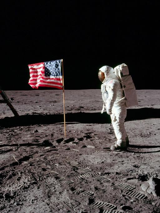 Historisches Foto, auf dem Neil Armstrong in seinem Raumanzug auf der Mondoberfläche steht und auf die US-amerikanische Flagge schaut.