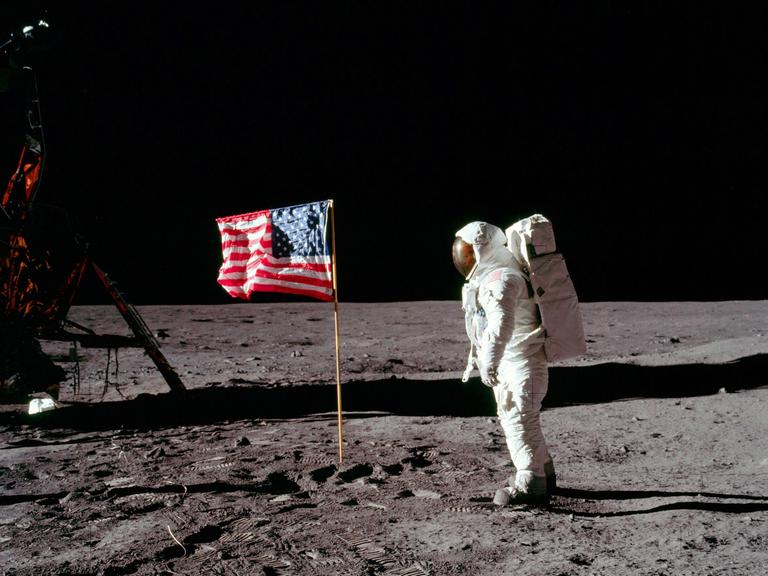 Historisches Foto, auf dem Neil Armstrong in seinem Raumanzug auf der Mondoberfläche steht und auf die US-amerikanische Flagge schaut.