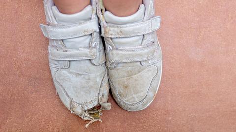 Kinderfüße in kaputten weißen Sportschuhen, die vorne ein Loch haben.