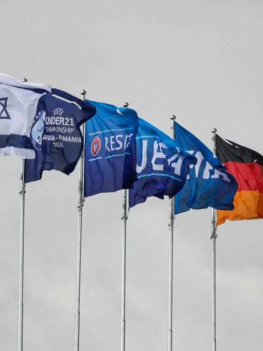Die Flaggen Israels, Deutschlands und der UEFA wehen am Stadion in Georgien. Im Hintergrund grauer Himmel.