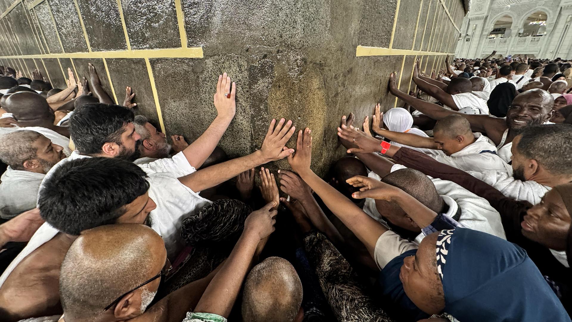 Viele Menschen drängen sich an die Steinwand der Kaaba in der Stadt Mekka. Sie halten die Hände gegen den Stein.