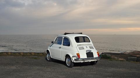 Ein alter, weißer Fiat steht am Meer, der Fahrer sitzt in dem Auto und blickt in Richtung Meer.