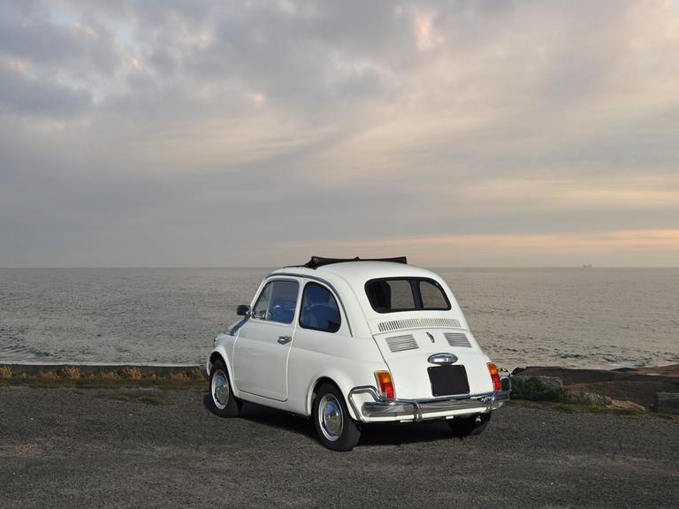 Ein alter, weißer Fiat steht am Meer, der Fahrer sitzt in dem Auto und blickt in Richtung Meer.