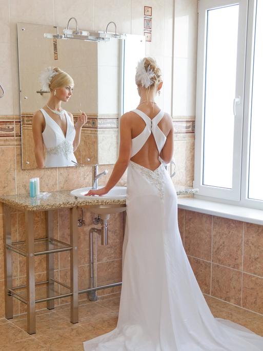 Eine Frau im weißen Brautkleid steht mit einem Lippenstift in der Hand vor dem Badezimmerspiegel.