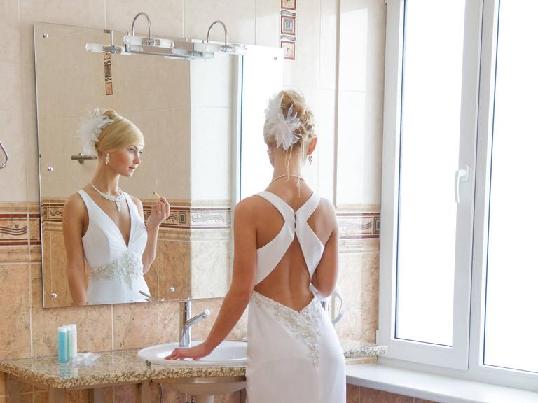 Eine Frau im weißen Brautkleid steht mit einem Lippenstift in der Hand vor dem Badezimmerspiegel.