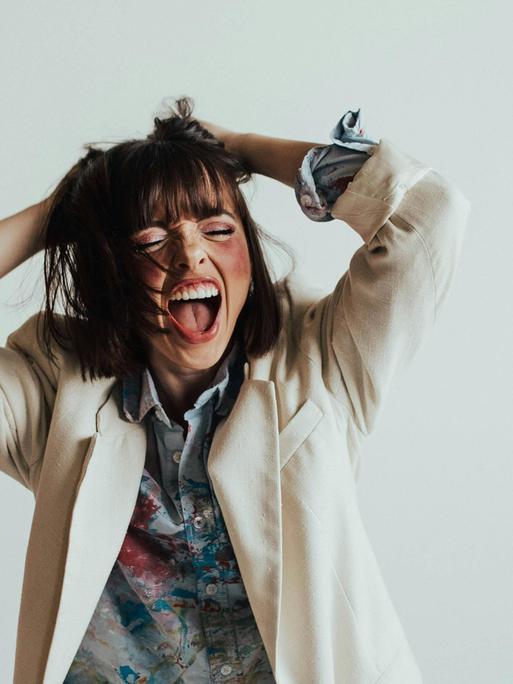 Eine junge Frau scheint laut zu singen mit weit offenem Mund und hebt dabei die Arme. 