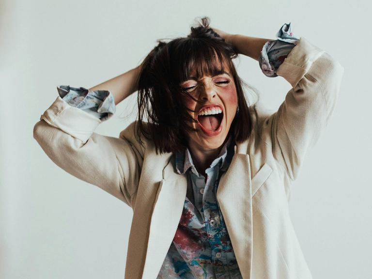 Eine junge Frau scheint laut zu singen mit weit offenem Mund und hebt dabei die Arme. 