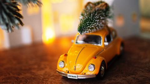 Auf einem kleinen, gelben Modellauto ist ein verschneiter Tannenbaum aufs Dach geschnallt und fährt durch die Modell-Schneelandschaft.