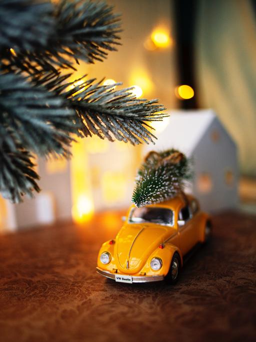 Auf einem kleinen, gelben Modellauto ist ein verschneiter Tannenbaum aufs Dach geschnallt und fährt durch die Modell-Schneelandschaft.