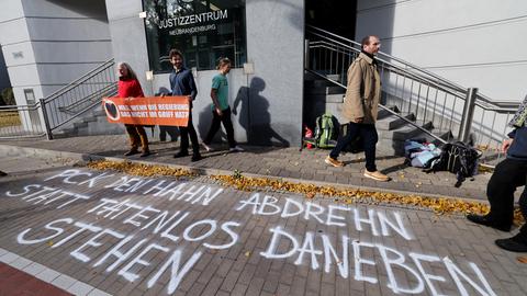  Mitglieder und Sympathisanten der Gruppe Letzte Generation stehen bei einer Mahnwache vor einem Gerichtsgebäude, in dem ein Zivilprozess stattfinden soll. Sie halten ein Transparent mit der Aufschrift "Was, wenn die Regierung das nicht im Griff hat?" steht.  Auf dem Bürgersteig im Vordergrund steht "PCK den Hahn abdrehn statt tatenlos daneben stehen". 