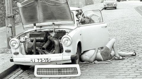 Zwei Menschen reparieren in Ostberlin einen Trabant. Die Aufnahme ist in Schwarz-weiß. Eine Person liegt unter dem Auto, eine andere werkelt an der Fahrerseite herum. 