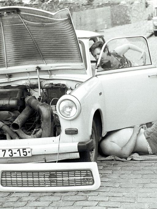 Zwei Menschen reparieren in Ostberlin einen Trabant. Die Aufnahme ist in Schwarz-weiß. Eine Person liegt unter dem Auto, eine andere werkelt an der Fahrerseite herum. 