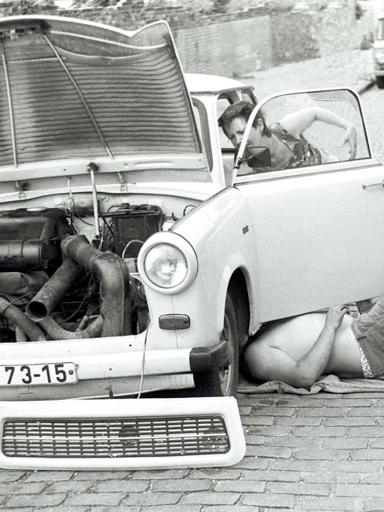Zwei Menschen reparieren in Ostberlin einen Trabant. Die Aufnahme ist in Schwarz-weiß. Eine Person liegt unter dem Auto, eine andere werkelt an der Fahrerseite herum. 