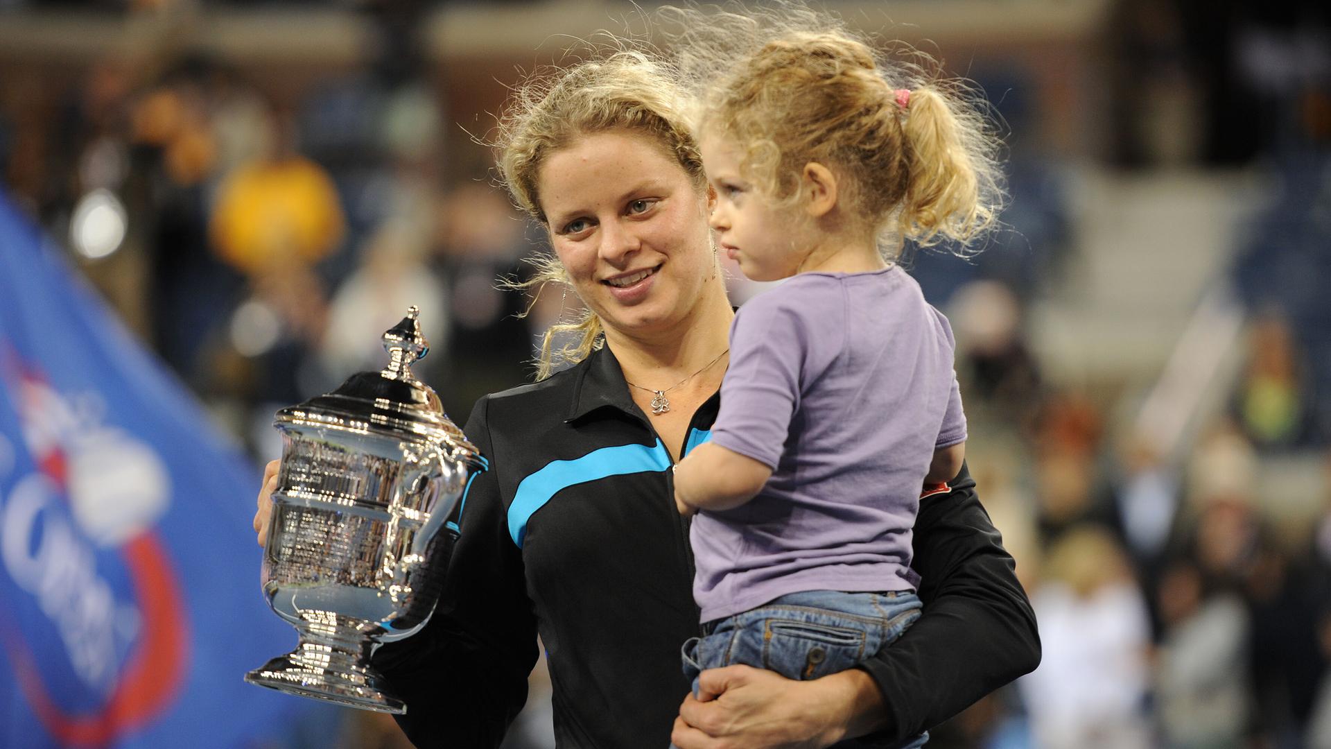 Klijsters in einer schwarzen Trainingsjacke hält mit der rechten Hand den silbernen Pokal und lächelt. Mit links hält sie ihre kleine Tochter im lilafarbenen T-Shirt auf dem Arm. Dahinter unscharf eine Zuschauertribüne.