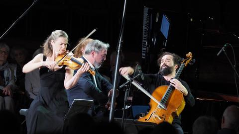 Ein Klaviertrio aus Geigerin, Cellist und Pianist spielt ein Konzert. Die Bühne ist ganz schwarz.