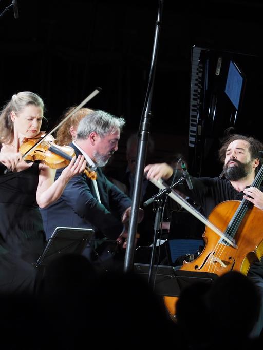 Ein Klaviertrio aus Geigerin, Cellist und Pianist spielt ein Konzert. Die Bühne ist ganz schwarz.