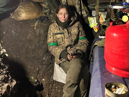 Eine junge blonde Frau sitzt in Soldatenuniform in einem niedrigen Erdbunker, sie sieht schmutzig und erschöpft aus und lächelt in die Kamera. 