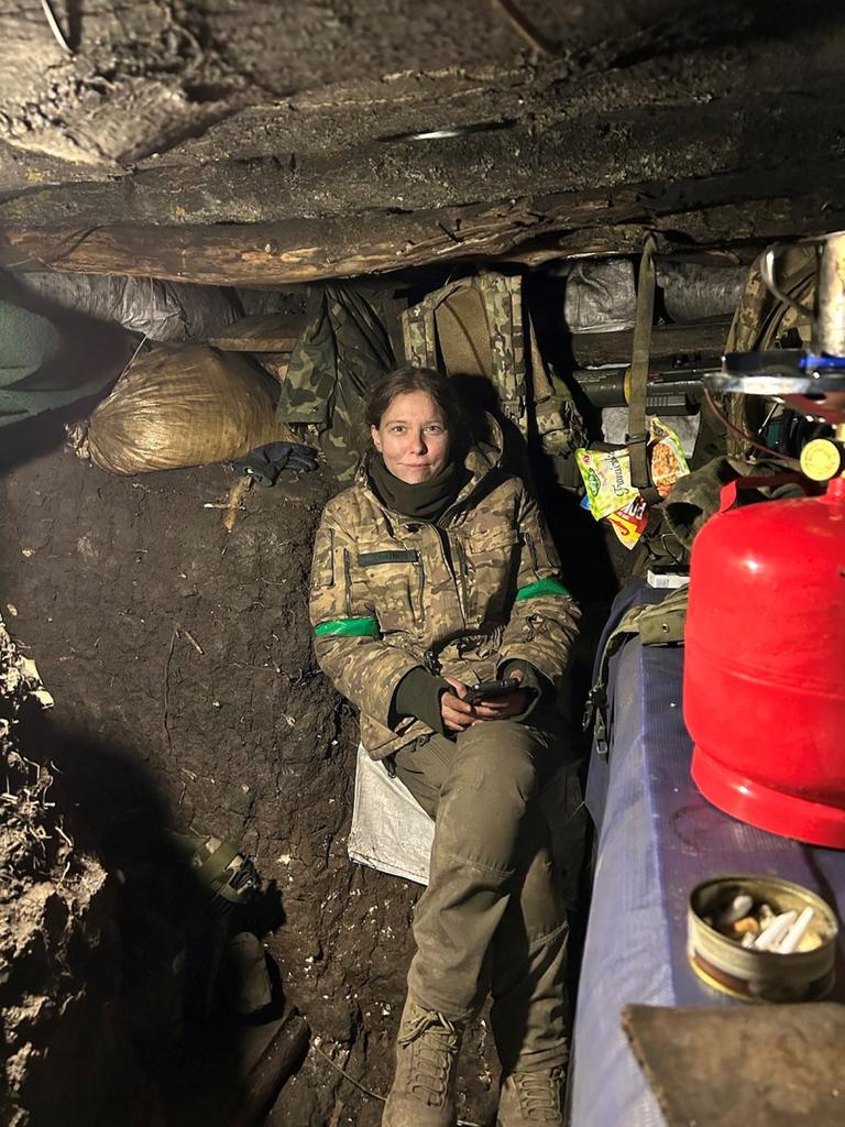 Eine junge blonde Frau sitzt in Soldatenuniform in einem niedrigen Erdbunker, sie sieht schmutzig und erschöpft aus und lächelt in die Kamera. 