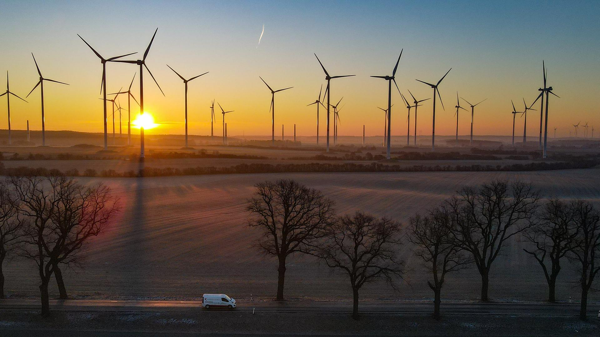 Im Hintergrund stehen Windräder vor dem Sonnenaufgang. Davor fährt auf einer Strasse ein Auto von links nach rechts.