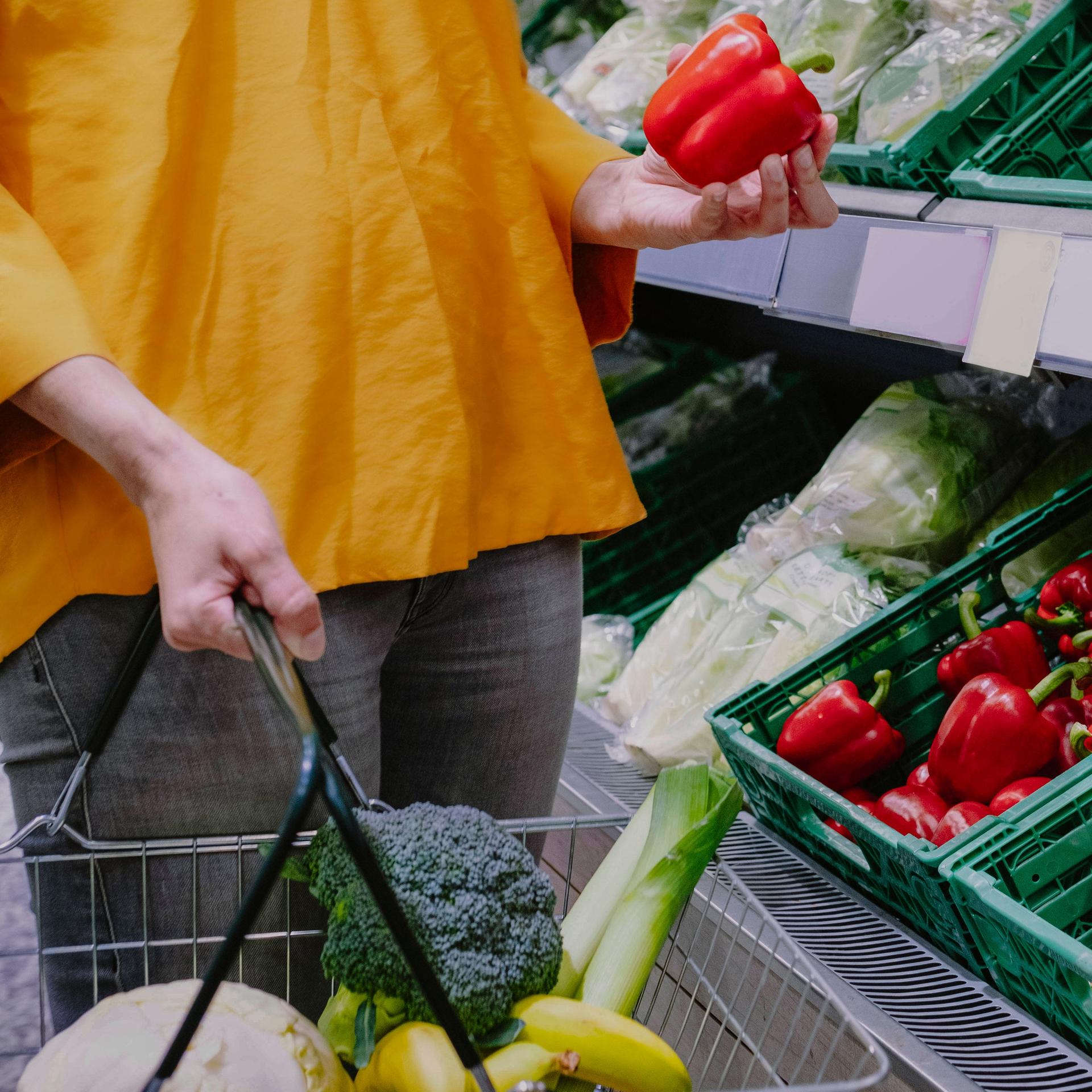 Eine Frau steht mit Einkaufskorb in einem Supermarkt vor dem Gemüseregal und hält eine rote Paprika in der Hand.