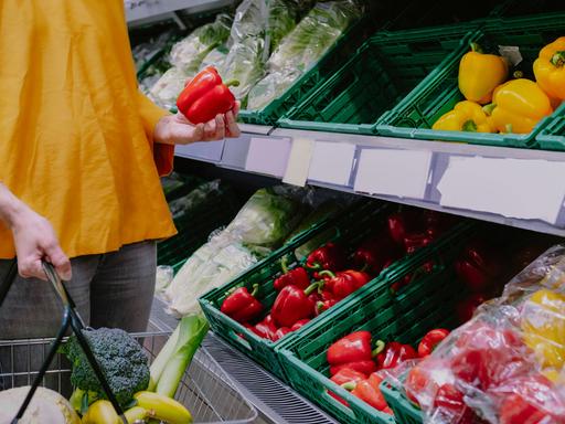Eine Frau steht mit Einkaufskorb in einem Supermarkt vor dem Gemüseregal und hält eine rote Paprika in der Hand.