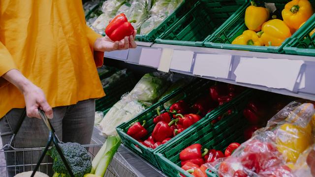 Eine Frau steht mit Einkaufskorb in einem Supermarkt vor dem Gemüseregal und hält eine rote Paprika in der Hand.