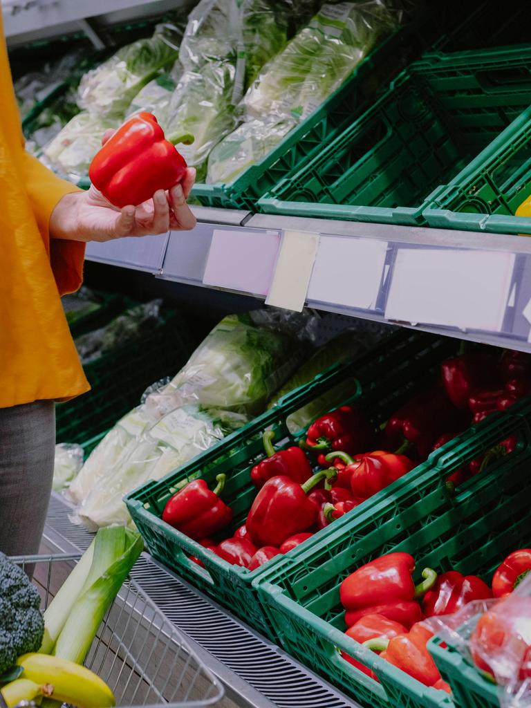 Eine Frau steht mit Einkaufskorb in einem Supermarkt vor dem Gemüseregal und hält eine rote Paprika in der Hand.