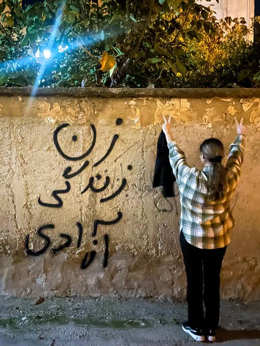 Eine junge iranische Frau ohne Hijab macht ein Peace-Zeichen und steht neben einer Wand, auf der auf Persisch Frau, Leben, Freiheit steht