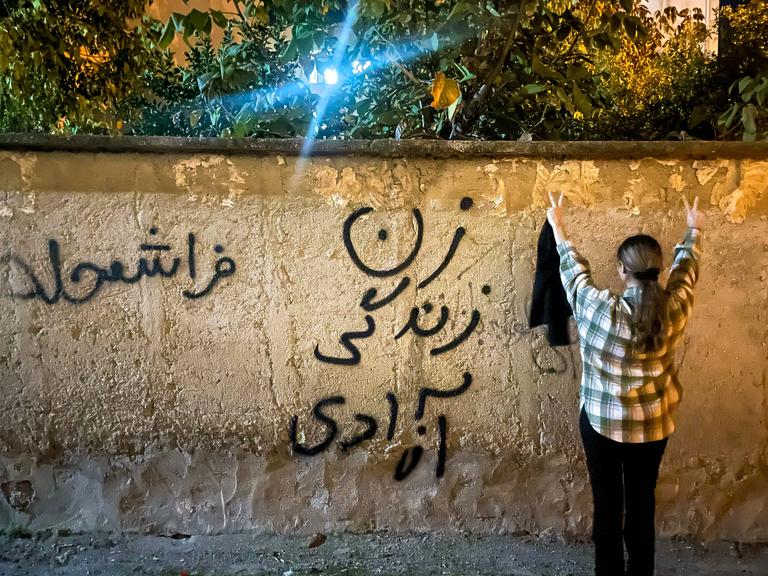Eine junge iranische Frau ohne Hijab macht ein Peace-Zeichen und steht neben einer Wand, auf der auf Persisch Frau, Leben, Freiheit steht