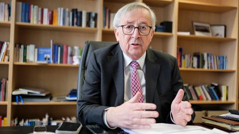 Der EU-Kommissionspräsident Jean-Claude Juncker sitzt vor einer Bücherwand in einem Büro an einem Arbeitstisch und spricht gestikulierend mit einem Gegenüber im Off 