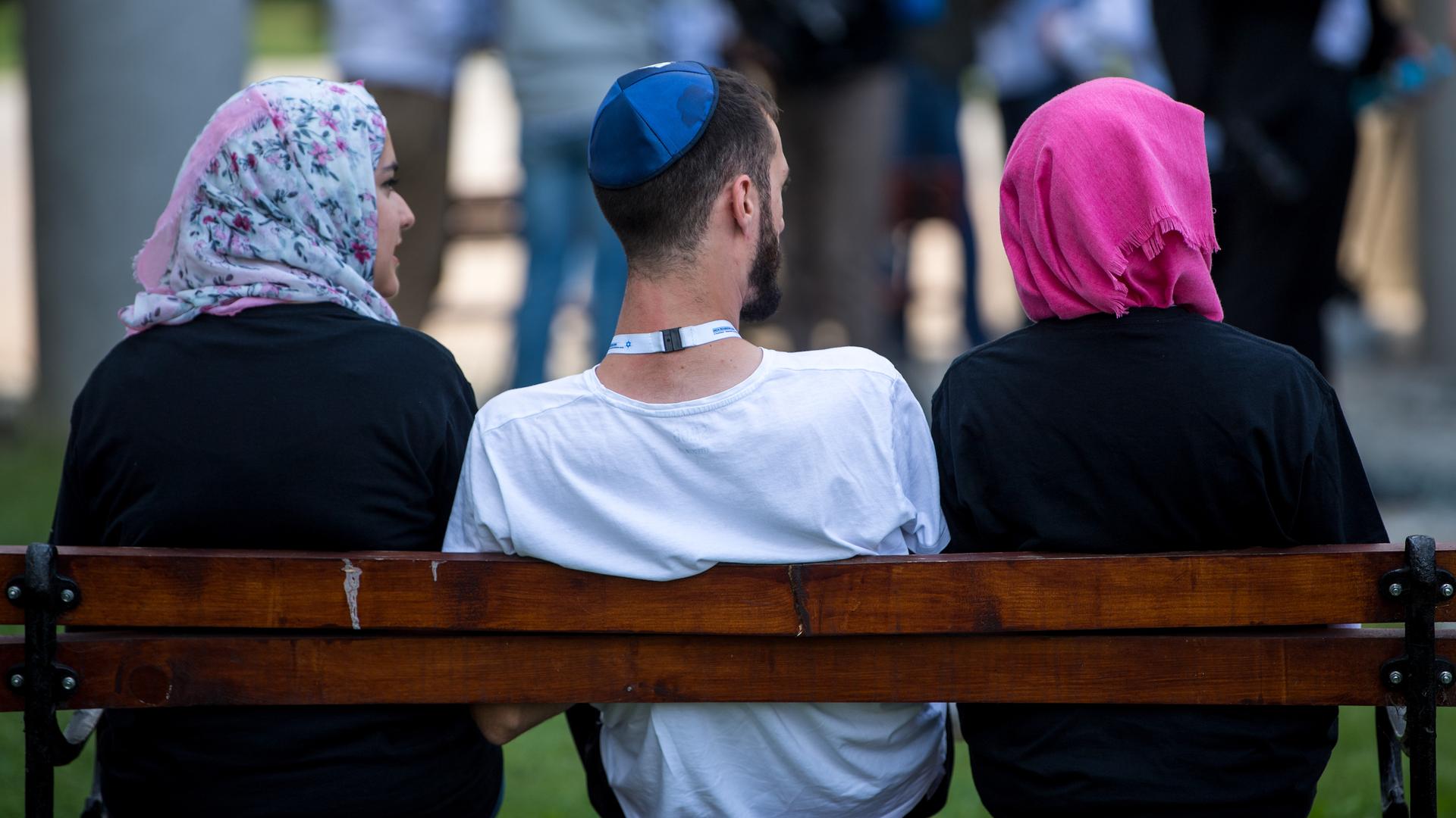 Ein jüdischer Mann mit Kippa sitzt zwischen zwei muslimischen Frauen auf einer Bank. Beide Frauen tragen Kopftuch.